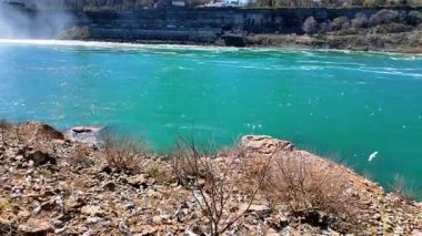 Ziyaretçiler, güneşli bir günde Niagara Şelalesi 'ndeki su akışını kayalık bir sahilden gözlemliyorlar. Ziyaretçiler Niagara Şelalesi 'nin kayalık kıyısında bekliyor.