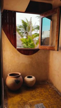 Pencere manzarası, basit bir odada palmiye ağaçları ve kil çömlekleri gösteriyor. Bir odanın içinde yerde iki kil çömlek var. Bir pencere açık havada dışarıda palmiye ağaçlarını gösterir.