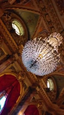 Altın yaldızlı tavana karşı kırmızı kadife ile süslü kristal avize. Büyük bir kristal avizenin alçak açılı görüntüsü, altın pervazlar ve dekore edilmiş bir tavandaki kırmızı örtü..