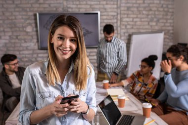 Meslektaşları arka planda mimari tasarımlar üzerinde çalışırken kendine güvenen genç kadın liderin portresi kameraya bakıyor..