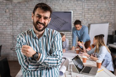 Meslektaşları arka planda mimari tasarımlar üzerinde çalışırken kendine güvenen genç erkek liderin portresi kameraya bakıyor..