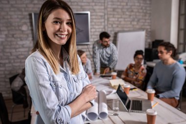 Meslektaşları arka planda mimari tasarımlar üzerinde çalışırken kendine güvenen genç kadın liderin portresi kameraya bakıyor..