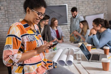 Meslektaşları arka planda mimari tasarımlar üzerinde çalışırken kendine güvenen genç kadın liderin portresi ayakta ve telefona bakıyor..