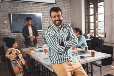 Meslektaşları arka planda mimari tasarımlar üzerinde çalışırken kendine güvenen genç erkek liderin portresi kameraya bakıyor.
