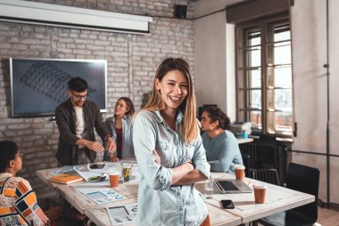 Meslektaşları arka planda mimari tasarımlar üzerinde çalışırken kendine güvenen genç kadın liderin portresi kameraya bakıyor..