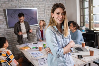 Meslektaşları arka planda mimari tasarımlar üzerinde çalışırken kendine güvenen genç kadın liderin portresi kameraya bakıyor..