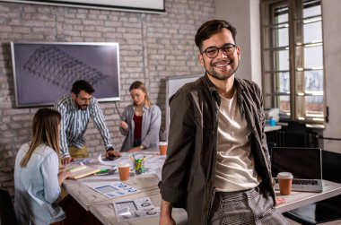 Meslektaşları arka planda mimari tasarımlar üzerinde çalışırken kendine güvenen genç erkek liderin portresi kameraya bakıyor..