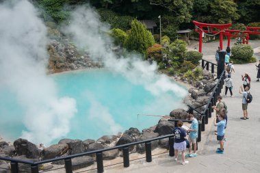 Japonya 'nın Oita, Beppu kentindeki Beppu kentinin arka planında kırmızı torii kapıları ile sudan yükselen buhar nedeniyle, turistler ana Umi Jigoku mavi kaplıcasını çevrelediler.