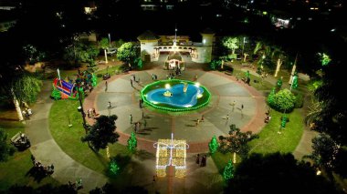 Kırsal kesimdeki küçük bir kasaba meydanında, insanların huzur içinde gezinmelerini sergiliyor, yemyeşil güzellikteki ağaçların ortasındaki hoş ışıklandırma..