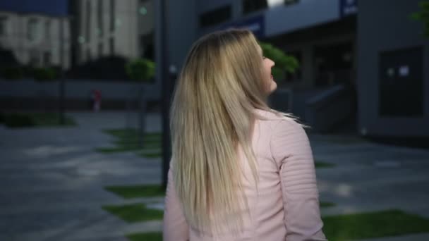 ピンクのジャケットを着たブロンドの女性。カメラに背を向けて立ちポーズ — ストック動画