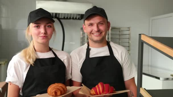 Portrait of two good looking young smiled male female bakers posing, croissants — Stock Video