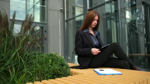 Jovem mulher trabalhando com tablet pc sentado em um banco no parque. Documentos femininos — Vídeo de Stock