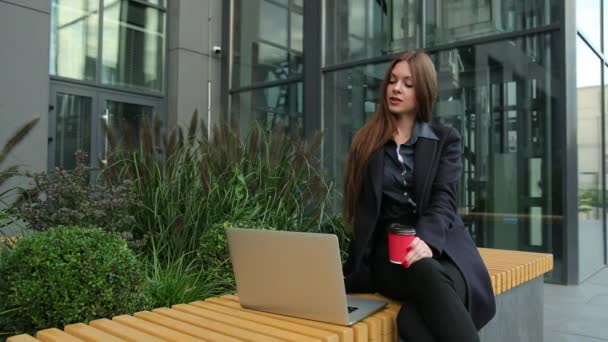 Mujer linda joven con portátil y café al aire libre en banco urbano de madera. — Vídeo de stock