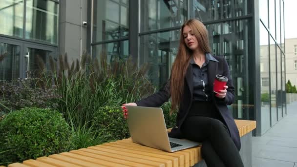 Jonge leuke vrouw met laptop en koffie buiten op houten stedelijke bank. — Stockvideo
