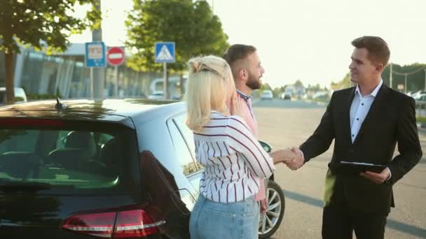 Vendedor vende el coche a la feliz pareja joven. Hombres estrechando las manos hermoso vehículo. — Vídeos de Stock