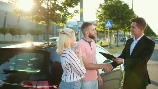 売り手は幸せな若いカップルに車を販売しています。購入者は車両購入に署名する — ストック動画