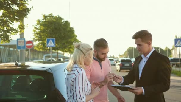 Vendedor vende el coche a la feliz pareja joven. Hombres estrechando las manos hermoso vehículo. — Vídeos de Stock