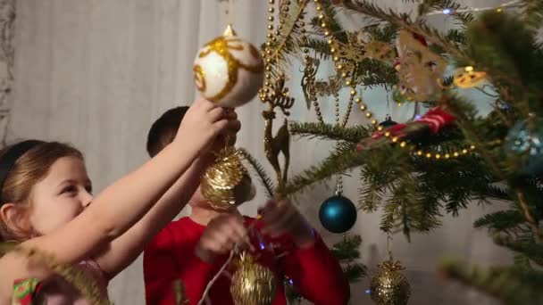 Украшение рождественской елки, вешание орнаментов, девочка и мальчик цепляются за игрушку — стоковое видео