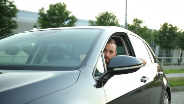 Un joven con barba mira por la ventana del coche, sonríe y levanta los pulgares — Vídeos de Stock