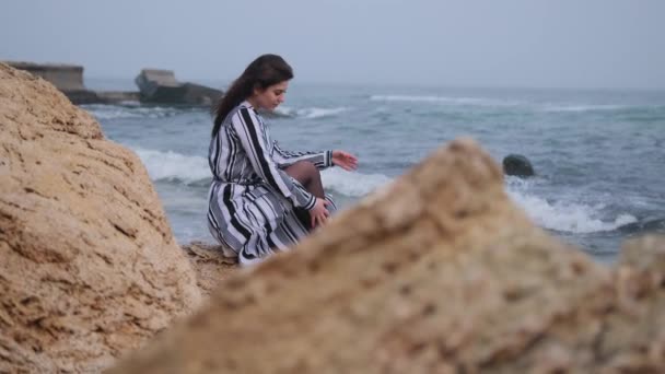 Νεαρή γυναίκα κάθεται στην κορυφή του βράχου, κοιτάζοντας την ακτή, βουνά. Τοπίο — Αρχείο Βίντεο