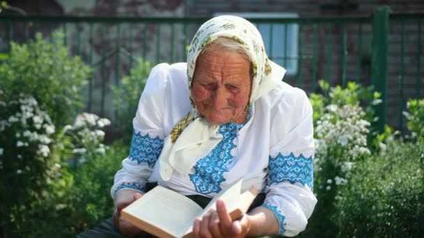 Una vieja abuela está sentada en la calle leyendo un libro. Vyshyvanka. — Vídeos de Stock