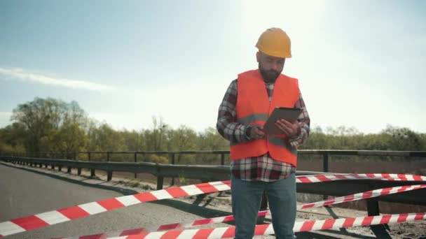 Jonge man met baard inspecteur in een oranje vest en beschermende helm met tablet — Stockvideo