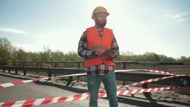 Jovem homem barbudo inspetor em um colete laranja e capacete protetor com tablet — Vídeo de Stock