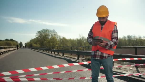 Jonge man met baard inspecteur in een oranje vest en beschermende helm met tablet — Stockvideo