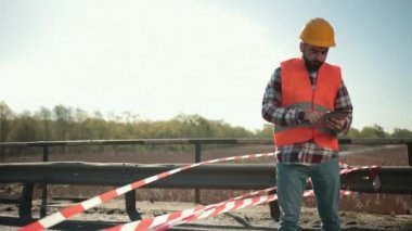 Turuncu yelek ve tabletli koruyucu kasklı genç sakallı müfettiş.