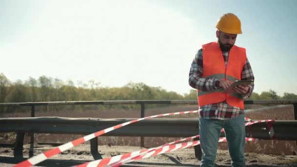 Jeune homme barbu inspecteur dans un gilet orange et casque de protection avec comprimé — Video