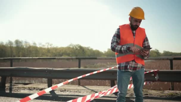 Mladý vousatý muž inspektor v oranžové vestě a ochranné přilbě s tabletem — Stock video