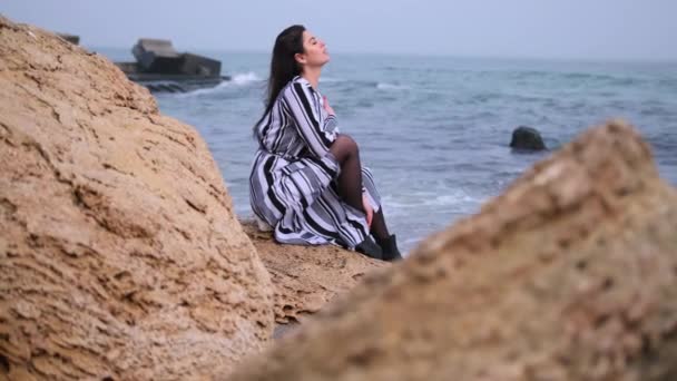 Женщина, сидящая на утесе с видом на океан с развевающимися волосами. Путешественница — стоковое видео