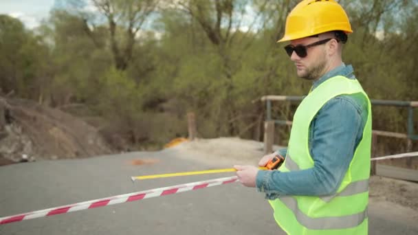 Szakállas mérnök védősisakos mérőszalaggal a kezében méri a távolságot — Stock videók