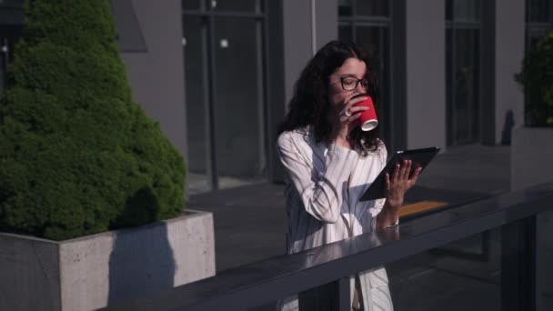 Νεαρή γυναίκα που χρησιμοποιεί ψηφιακό δισκίο έξω, χρησιμοποιώντας ψηφιακό δισκίο, πίνοντας καφέ — Αρχείο Βίντεο