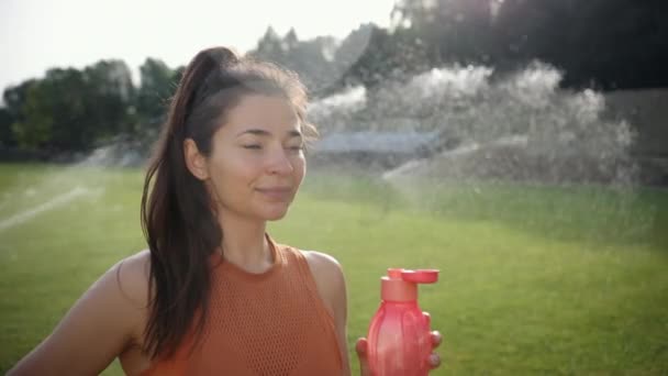 Mulher esportiva jovem bonita bebe água de garrafa de plástico, têmporas — Vídeo de Stock