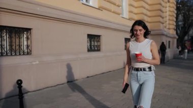 Cep telefonu konuşması, volta atma, kahve fincanı. Beyaz bluzlu ve kot pantolonlu genç bir kadın şehir caddesinde yürüyor, telefon konuşmaları yapıyor ve elinde kırmızı fincan kahve tutuyor..