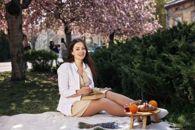Bahar sahnesi, piknik düzeni, mevsimlik meyveler. Çiçek açan kiraz ağaçlarının altında piknik battaniyesinde oturan genç kadın, kitaptan zevk alıyor ve yanında atıştırmalıklar ve meyvelerle çiçek tutuyor..