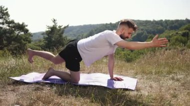 Mor minder, yoga seansı, doğanın farkındalığı. Doğanın kuşattığı çimen tarlasında, genç adam mor şort ve beyaz tişört giymiş, mor minder üzerinde yoga pozu veriyor..