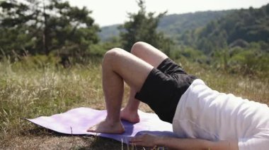 Yoga dengesi, yoga sağlığı, barışçıl doğa. Doğanın güzellikleriyle çevrili genç adam yoga dengesi kuruyor mor paspasın üzerinde, beyaz tişört ve siyah şort giyiyor..