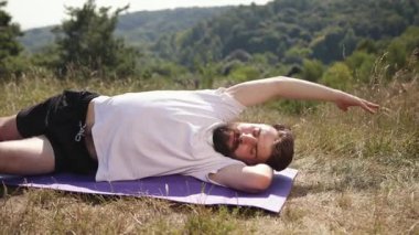 Yoga, kişisel bakım, genç adam, doğa bağlantısı. Beyaz tişörtlü ve siyah şortlu genç adam, doğa dolu çimenlik alanda mor paspas üzerinde yoga yaparak farkındalık kazanıyor..