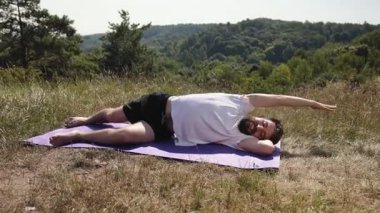 Yoga dengesi, yoga gücü, doğa bağlantısı. Genç adam, çimenli çayırlarda mor yoga minderinde, beyaz tişört ve siyah şort giyip, doğa ile çevrili farkındalığı ve dengeyi kucaklıyor..