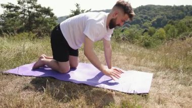 Denge pozisyonu, genç adam, yoga yolculuğu. Beyaz t-shirt ve siyah şort giyen genç adam, çimenli çayırlarda mor paspasın üzerinde yoga pozu vererek doğayı kucaklıyor..