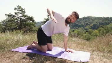 Yoga çalışması, yoga sükuneti, yoga sahası. Çimen tarlasında mor hasırda, genç adam yoga pozunu dengeleyerek farkındalık uyguluyor, siyah şort ve beyaz tişört giyiyor, doğayla bağlantılı..