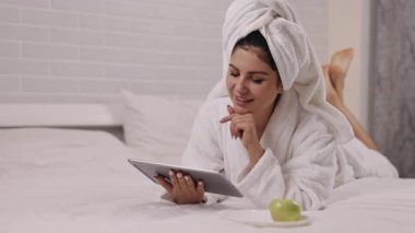 Sağlıklı hoşgörü, kadın dinlenmesi, ev eğlencesi. Kadın yatak odasında kişisel zaman geçirmeyi, karnına yaslanmayı, tablet üzerinde gezinmeyi ve elma tatmayı seviyor..