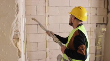 Alçı, sprey sıva, inşaat aletleri. Sert şapka ve güvenlik yeleği giyen inşaat işçisi yenileme sırasında duvara sprey tabancayla çimento harç sıvamış..