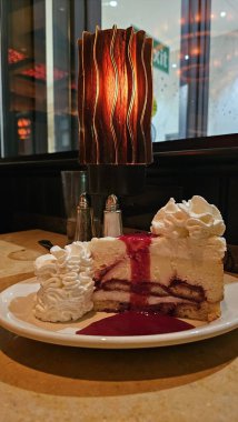 Krem şantili ve dut soslu dilimlenmiş çilekli kek sıcak ortam ışıklandırmalı sıcak bir restoran ortamında servis edilir hoşgörülü bir tatlı sunumu ve kafein yemek atmosferi vurgulanır..