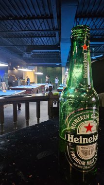 Bilardo salonundaki bir bar tezgahında Heineken bira şişesi. Arka planda bilardo masaları ve oyuncular. Kapalı bir eğlence mekanında gece hayatı, eğlence ve markalı içecek tüketimini gösteriyor. 2 Aralık 2024