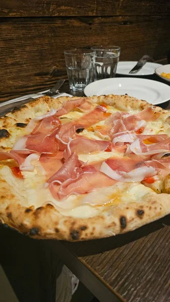 Eritilmiş peynirle kaplı ahşap kaplı İtalyan pizzasının yakın plan çekimi ve rustik bir restoran masasında jambon dilimleri, sanatsal mutfağı ve sıcak yemek ortamını ön plana çıkarıyor..