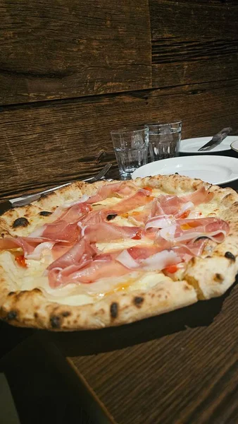 Eritilmiş peynirle kaplanmış ahşap bir masada servis edilen ince dilim jambonlu İtalyan pizzası geleneksel mutfak ve sıcak yemek atmosferini ön plana çıkarıyor..