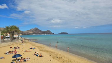 Portekiz 'in Madeira kentindeki Porto Santo plajının panoramik manzarası, altın kum, turkuaz Atlantik suyu, rahat sahil ziyaretçileri, palmiye çizgili gezinti güvertesi ve dokulu bulutların altındaki volkanik ada manzarası..